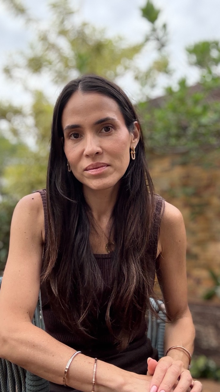 Dani Hayoshi sentada ao ar livre, com cabelo longo escuro e blusa escura, diante de árvores.